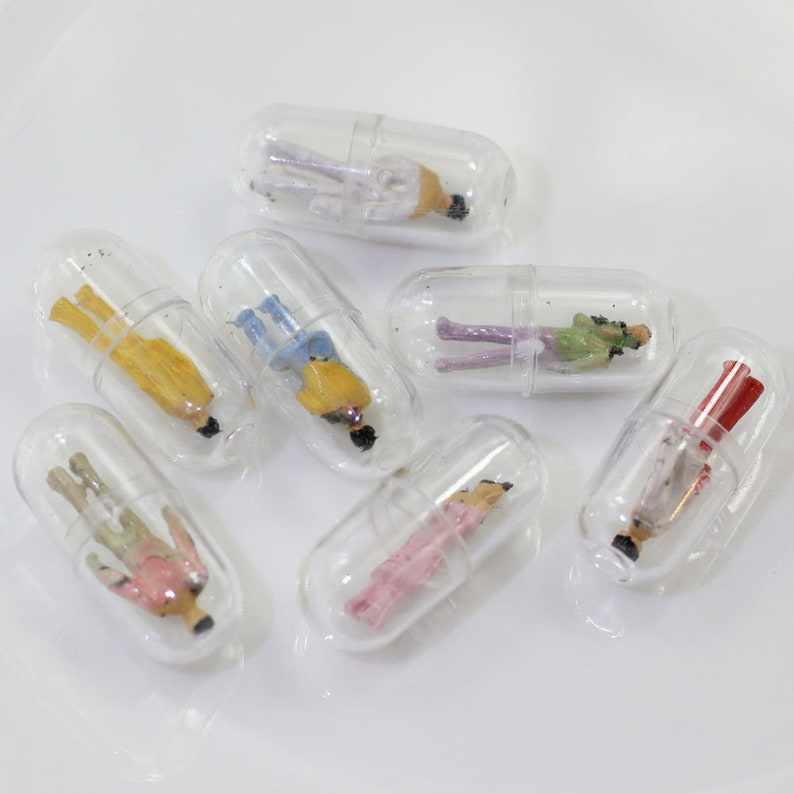 Op de afbeelding: Acht transparante plastic capsules, elk met een klein beeldje van een persoon in een andere gekleurde outfit. De beeldjes zijn gerangschikt op een wit oppervlak.