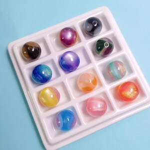 May include: A white plastic tray with twelve square compartments, each containing a single spherical bead. The beads feature various colors including blue, yellow, pink, orange, purple, and green, with some displaying glittery or marbled finishes.