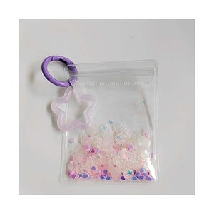 Puede incluir: Una bolsa rectangular transparente con cierre de cremallera, llena de lentejuelas rosas y blancas y confeti en forma de estrella. Un mosquetón morado y un dije de estrella translúcida están unidos a la bolsa.