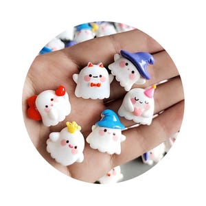 Peut inclure: Une collection de petits charmes blancs en forme de fantômes avec divers accessoires colorés. Certains fantômes portent des chapeaux, des nœuds ou des couronnes. Les charmes ont des joues roses et sont probablement destinés à l'artisanat ou à la bijouterie.
