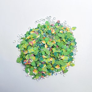May include: A collection of small, flat, decorative polymer clay slices in the shapes of green leaves, yellow and orange fish, and green frogs, mixed with tiny, clear, round, faceted acrylic rhinestones scattered throughout the pile.