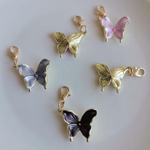 May include: Five butterfly charms in gold, black, gray, and pink with gold-tone clasps. The butterfly wings have a metallic sheen. The charms are arranged on a white surface. These charms are suitable for jewelry making or craft projects.