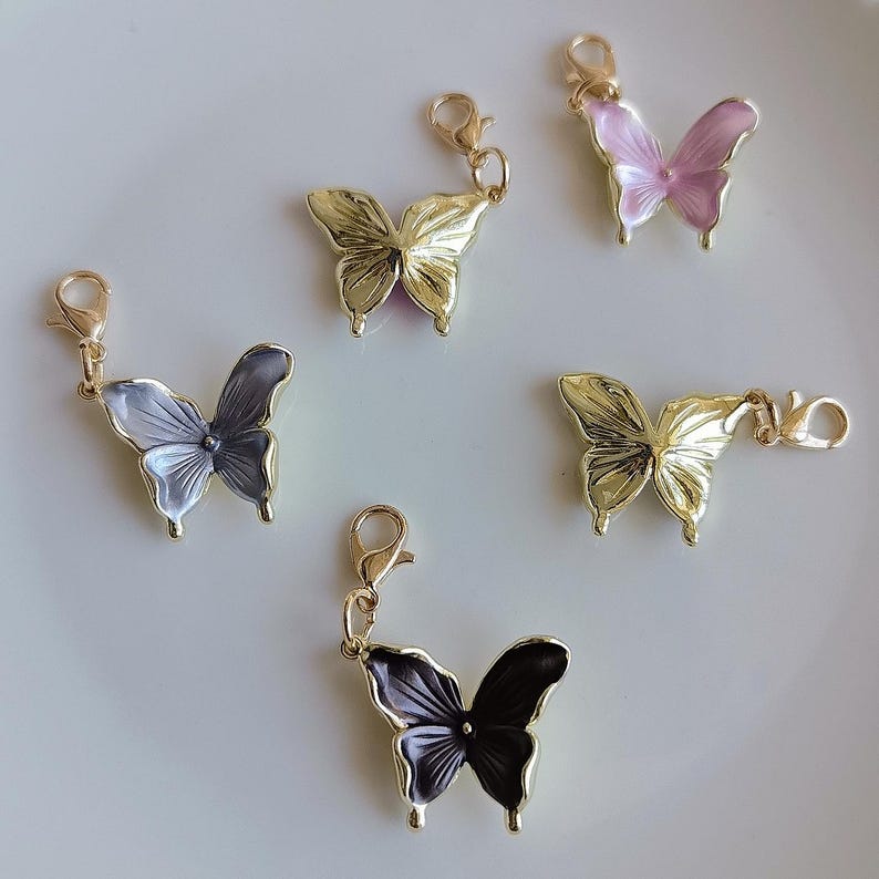 May include: Five butterfly charms in gold, black, gray, and pink with gold-tone clasps. The butterfly wings have a metallic sheen. The charms are arranged on a white surface. These charms are suitable for jewelry making or craft projects.