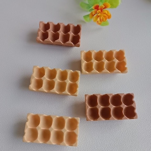 May include: Five miniature egg cartons in varying shades of brown and beige are arranged on a white surface. Each carton has a rectangular shape with twelve rounded egg compartments. A small flower with yellow and orange petals is in the background.