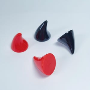 May include: Four small, smooth, conical horn-shaped objects with a glossy finish. Two of the horns are bright red and two are solid black. Each piece features a curved, pointed tip and a flat, circular base, arranged in a scattered formation.