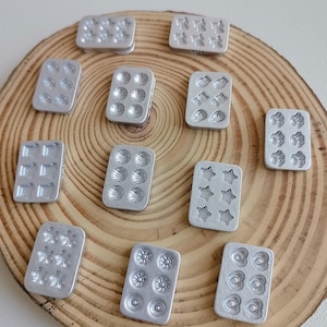 May include: A collection of small, silver-colored baking molds in various shapes, including stars, hearts, flowers, and shells. The molds are arranged on a wooden surface, showcasing their detailed designs and potential for creating miniature treats.
