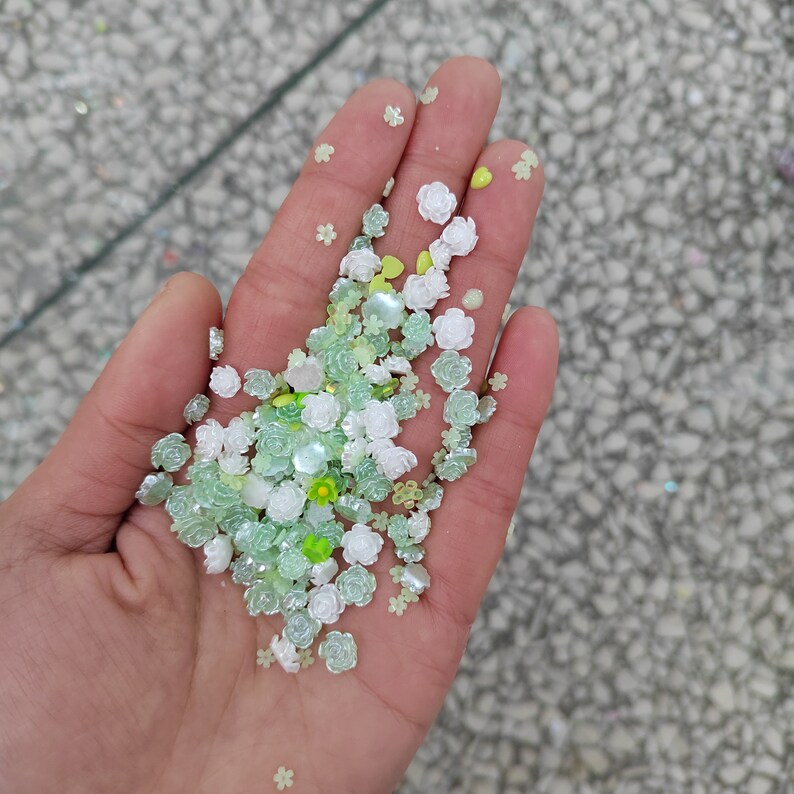 May include: A hand holding a variety of small, green and white flower-shaped embellishments. The embellishments are scattered on the palm of the hand and are made of plastic or resin.