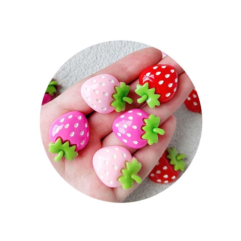 May include: A collection of colorful strawberry-shaped embellishments. The strawberries are in shades of pink and red, with green leafy tops. The embellishments have a glossy finish and are scattered across a surface.