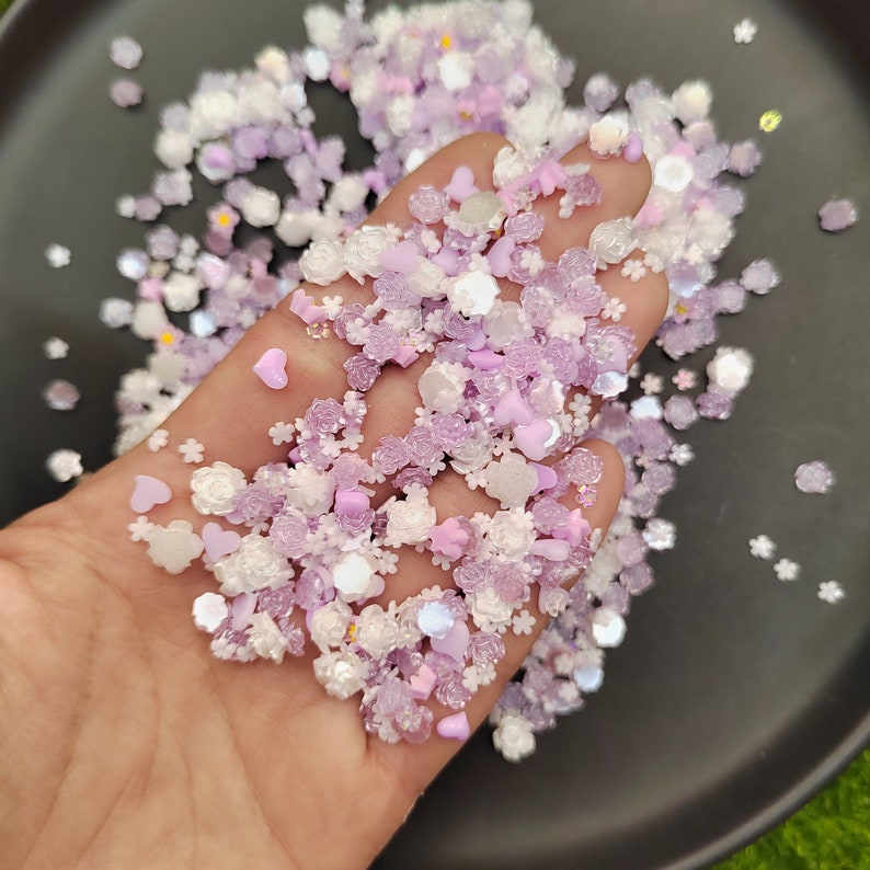 May include: A mix of white and purple resin flower and heart shaped sprinkles. The sprinkles are scattered on a black surface.