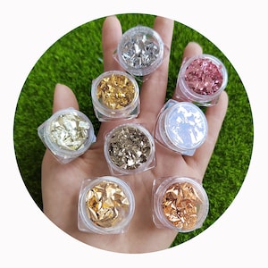May include: Seven small jars of metallic flakes in various colors, including gold, silver, and pink. The jars are arranged in a circle on a hand.