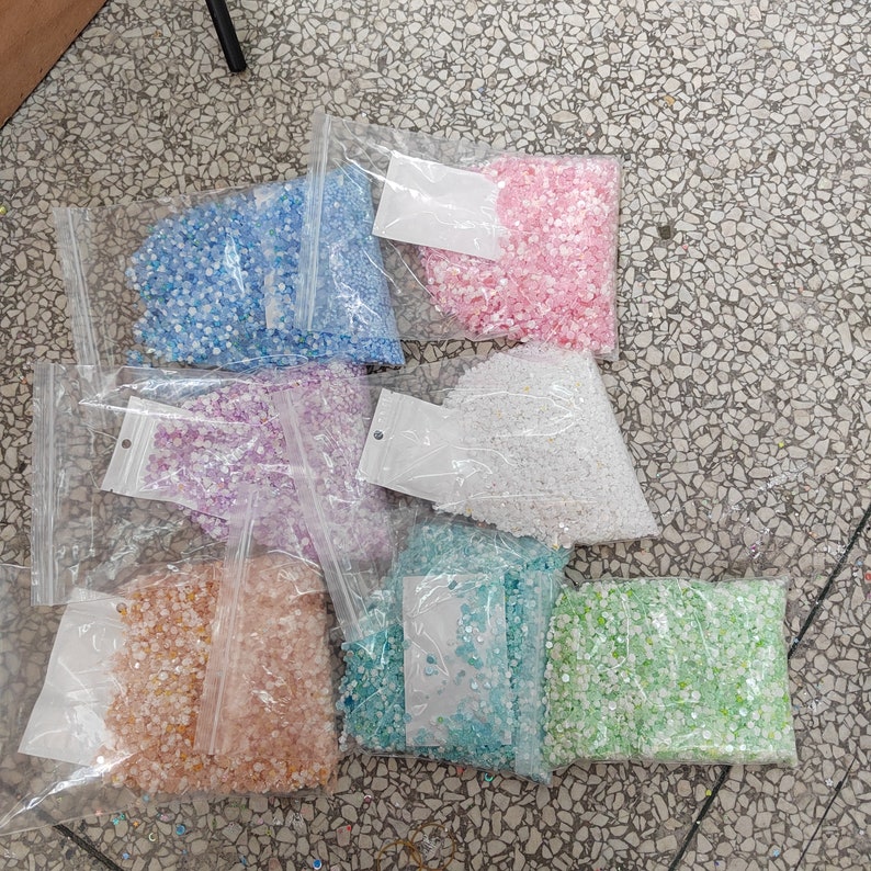 May include: Seven bags of iridescent sequins in various colors, including pink, blue, green, white, and purple. The sequins are packaged in clear plastic bags with white labels.