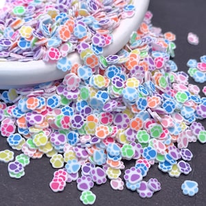 Puede incluir: Un montón de rodajas de arcilla polimérica con forma de huellas de patas multicolores. Los colores incluyen rosa, morado, azul, verde, amarillo y naranja. Las piezas de arcilla están esparcidas alrededor de un cuenco blanco, perfectas para nail art u otros proyectos de manualidades.
