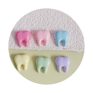 Puede incluir: Seis amuletos de resina en forma de diente de color pastel en rosa, azul claro, amarillo, verde, morado y rosa. Los amuletos están dispuestos en una fila sobre una superficie blanca y verde.