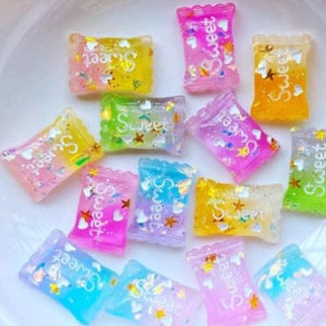 May include: Assortment of colorful, glittery candy charms with "Sweet" written on each one. The charms are shaped like small, rectangular candy bags.