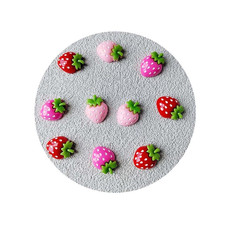 May include: A collection of strawberry-shaped embellishments in various shades of pink and red, each with green leafy tops. The strawberries are arranged on a textured, gray circular surface, creating a playful and decorative design.