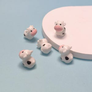 May include: Five small, white cow figurines with pink noses and black spots are scattered on a light blue surface. The figurines are ceramic and have a glossy finish. One figurine is upright, while the others are in various poses.