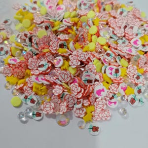 Puede incluir: Primer plano de una colección de materiales de manualidades. La imagen muestra decoraciones en forma de rosa rojas y blancas, piezas amarillas en forma de estrella, lentejuelas iridiscentes transparentes y pequeños trozos amarillos redondos. Los artículos están esparcidos sobre una superficie blanca.