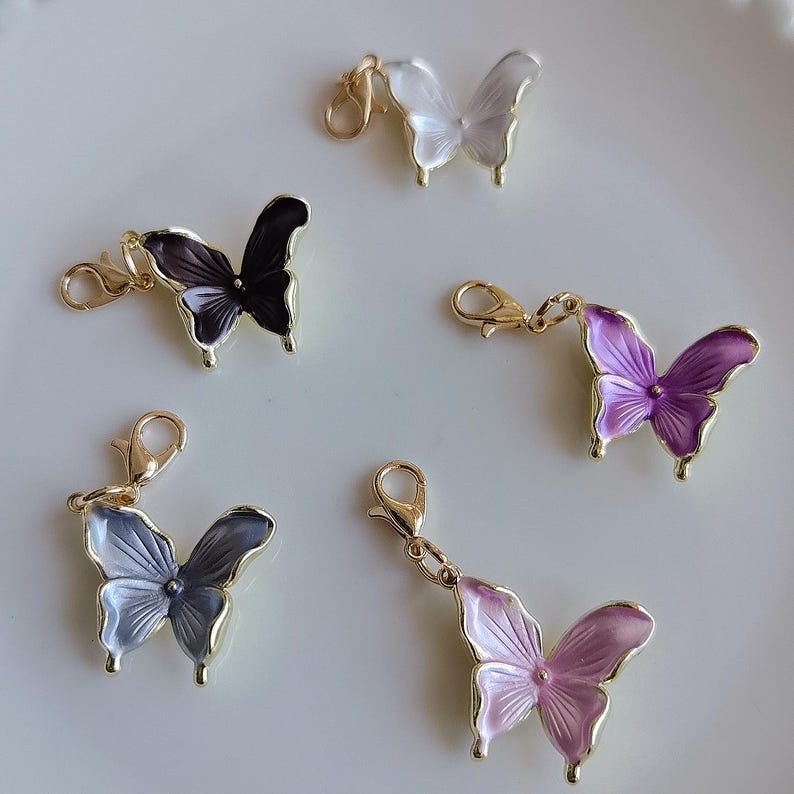 May include: Five butterfly charms in various colors: white, black, purple, lavender, and pink. Each charm has a gold-colored clasp and a gold-colored outline. The butterfly wings are detailed with a glossy finish, and the charms are arranged on a white surface.