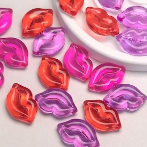 Könnte beinhalten: Eine Sammlung transluzenter Lippen-förmiger Perlen in Rot-, Rosa- und Lilatönen. Die Perlen sind auf einer weißen Oberfläche verstreut, einige befinden sich auf einem weißen Teller. Die glänzende Oberfläche der Perlen reflektiert das Licht.