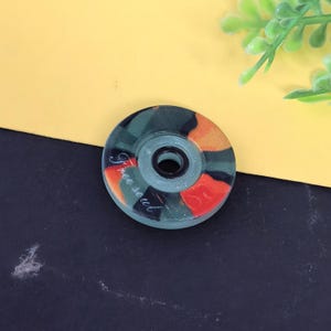 May include: A round, decorative bottle stopper with a central hole. The stopper features a camouflage pattern in shades of green, orange, and red. The words "Free soul" are written in cursive script on the surface.