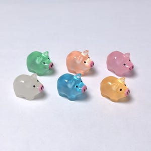 Puede incluir: Seis figuritas de cerdos en miniatura de varios colores: verde, melocotón, rosa, blanco, azul y amarillo. Cada cerdo tiene un hocico rosa y pequeños ojos negros. Las figuritas están dispuestas sobre una superficie blanca.