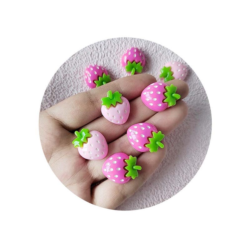 May include: A collection of small, strawberry-shaped embellishments. The strawberries are pink and fuchsia with green stems. Some have white dots. The items are arranged on a light pink surface.