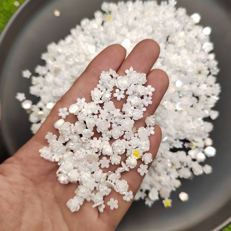 May include: A hand holding a large amount of white flower-shaped embellishments. The embellishments are made of plastic and have a pearlescent finish.