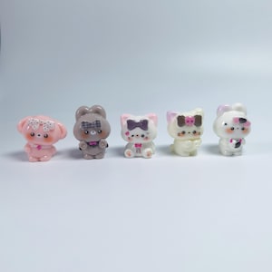 Peut inclure: Cinq petites figurines d'animaux alignées. De gauche à droite : un ours rose avec un nœud à pois, un ours gris avec un nœud à carreaux, un chat blanc avec un nœud foncé, un chat blanc avec un nœud tête de mort et un lapin blanc avec un nœud rose.