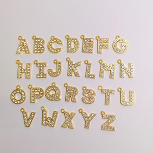 Könnte beinhalten: Eine Sammlung von sechsundzwanzig goldfarbenen Metall-Buchstabenanhängern, die jeweils mit einer Reihe von klaren, funkelnden Strasssteinen besetzt sind. Jeder Buchstabe von A bis Z ist enthalten, und jeder Anhänger hat eine kleine Öse oben.