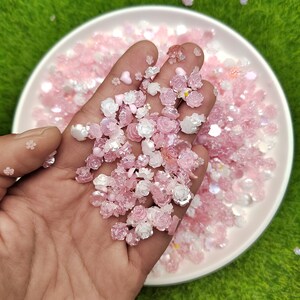 May include: A close-up of a hand holding a variety of pink and white resin flower embellishments. The embellishments are in a bowl and are surrounded by other pink and white embellishments.