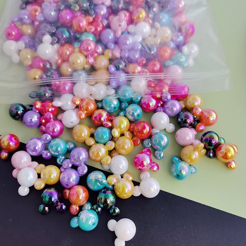 K&ouml;nnte beinhalten: Eine gro&szlig;e Auswahl an schillernden Perlen in Mickey-Maus-Kopfform in verschiedenen Farben, darunter Pink, Lila, Blau, Gr&uuml;n, Gelb und Orange. Die Perlen sind auf einer gr&uuml;n-schwarzen Oberfl&auml;che verstreut.