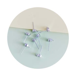 Peut inclure: Des attaches de boucles d'oreilles en métal argenté avec une boucle en haut et un petit capuchon rond en bas. Les attaches sont disposées sur une surface vert clair.
