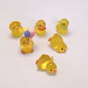 Puede incluir: Colección de seis pequeñas figuras de patos amarillos translúcidos. Algunos patos tienen detalles rojos o azules, y uno sostiene globos. Los patos están en varias poses, incluyendo de pie, sentados y tumbados. Estas figuras en miniatura son probablemente para manualidades o decoración.