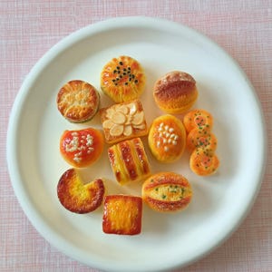 May include: A white plate displays an assortment of miniature baked goods. The collection includes round pastries with seeds, almond-topped squares, and various other small, colorful treats. The arrangement is set against a pink background.