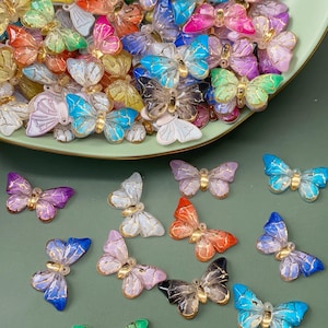 Puede incluir: Una colección de adornos decorativos con forma de mariposa. Las mariposas son de varios colores, incluyendo rosa, azul, verde y amarillo, con detalles dorados. Algunas están esparcidas sobre una superficie verde, otras en un plato verde.