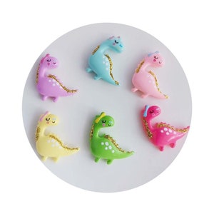 Puede incluir: Seis figuritas de dinosaurio de colores con detalles de purpurina. Los dinosaurios son de varios colores, incluyendo rosa, azul, verde y amarillo. Cada dinosaurio tiene una cara linda con los ojos cerrados y un pequeño lazo en la cabeza.