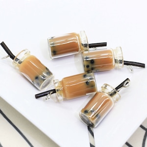 May include: Five clear glass charms shaped like boba tea cups with black straws and brown liquid inside. The charms have black boba pearls inside the liquid.