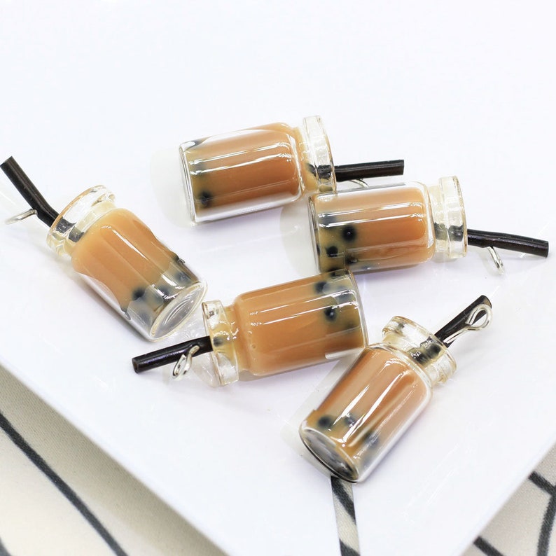 May include: Five clear glass charms shaped like boba tea cups with black straws and brown liquid inside. The charms have black boba pearls inside the liquid.