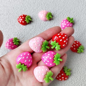 Op de afbeelding: Een verzameling aardbeivormige versieringen in verschillende tinten roze en rood. Elke aardbei heeft een groene steel en is bezaaid met witte stippen. De versieringen zijn verspreid over een lichtgrijze ondergrond.