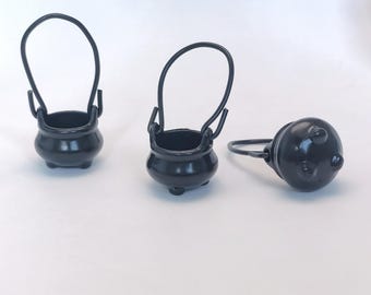 Op de afbeelding: Drie kleine, zwarte metalen ketels met gebogen handvatten. Elke ketel heeft een afgeronde body en drie kleine voetjes. Eén ketel ligt op zijn kant. De ketels zijn waarschijnlijk decoratieve items, mogelijk voor Halloween of fantasy-thema knutselen.