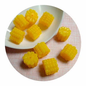 May include: Eight yellow corn on the cob shaped resin beads. The beads are arranged on a white plate and a pink and white patterned surface.