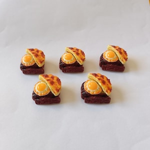 Könnte beinhalten: Fünf Miniatur-Lebensmittel, jeweils bestehend aus einem Spiegelei mit gelbem Eigelb und einer Scheibe goldbraunem Brot, die auf einem dunkelbraunen Patty liegen. Anordnung auf einer weißen Oberfläche.
