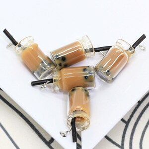 May include: Five clear glass bottles with brown liquid and black straws. The bottles have black boba pearls inside. The bottles are arranged in a cluster on a white surface.