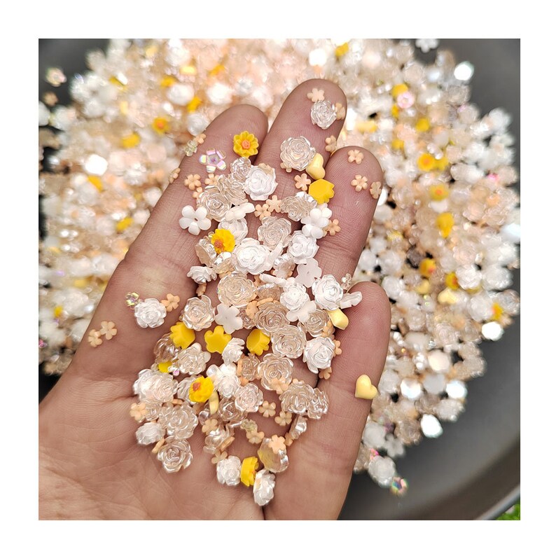 May include: A handful of small, colorful, plastic flower and heart shaped sprinkles. The sprinkles are in shades of white, yellow, and orange.