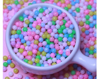 May include: A white ceramic bowl filled with pastel-colored round sprinkles. The sprinkles are in shades of pink, blue, green, and yellow.