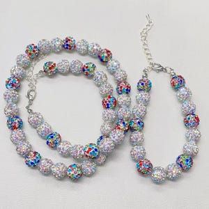 Può includere: Un set di due collane con perline di cristallo colorate. La collana più grande è più lunga e ha una chiusura argentata. La collana più piccola è più corta e ha una chiusura argentata. Entrambe le collane sono fatte di perline bianche con cristalli colorati.