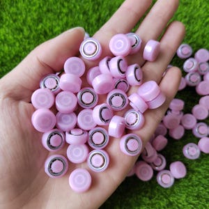 Può includere: Primo piano di una mano che tiene una grande quantità di piccoli tappi rotondi di plastica rosa con un anello argentato al centro. I tappi hanno una finitura lucida e lucida.