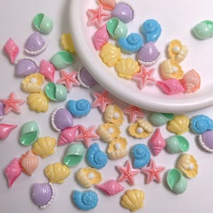 Puede incluir: Una colección de pequeños cabujones de resina de colores con formas de conchas marinas, estrellas de mar y almejas con perlas. Las piezas son de tonos pastel rosa, azul, amarillo, verde y morado, esparcidas sobre una superficie blanca.