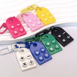May include: A collection of colorful calculator keychains in various colors, including pink, yellow, green, black, blue, and white. Each keychain features a rectangular design with raised calculator buttons and a metal clasp for attachment.