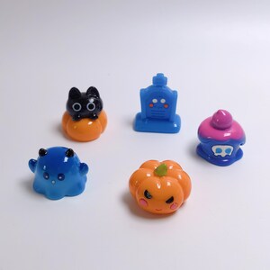 May include: A set of five small, glossy, colorful figurines including a black cat on an orange pumpkin, a blue tombstone, a blue ghost with black horns, an orange pumpkin with a green stem, and a blue cupcake with a pink top and skull icon.